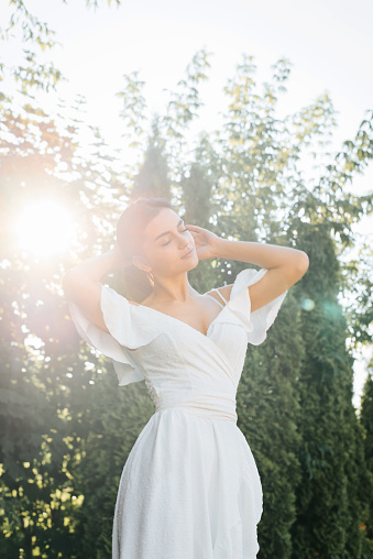Elegant young brunette woman posing with closed eyes in sunbeam, beautiful smiling lady with dark skin in white summer dress enjoys, outdoors. Beauty and fashion, vertical photo.