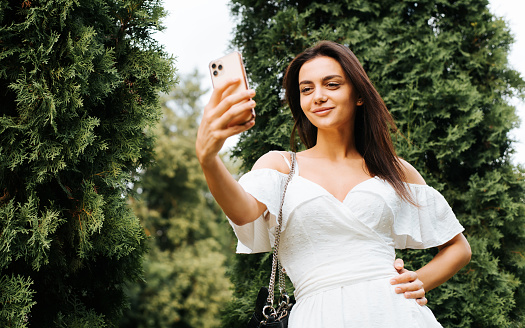 Beautiful young woman in white light dress smiling pleasantly at phone camera, taking positive selfie on smartphone, posing for photo in park outdoors. Elegant indian woman summer lifestyle.