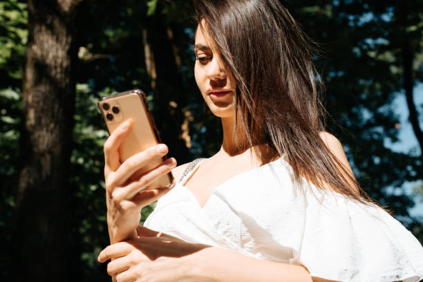 istock Young brunette woman in white dress using smartphone, chatting with someone, browsing internet on mobile phone outdoors. Beautiful girl holding phone, low angle view, selective focus on woman's face 1332339104