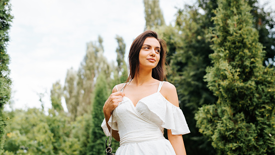 Low angle view portrait of pretty young woman wearing white dress and handbag posing in park outdoors and looking to the side, lifestyle. Smiling girl model, beauty and fashion concept.