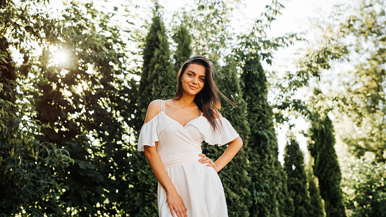 Fashionable young brunette woman elegantly posing in white summer dress outdoors looking cute at camera, low angle. Smiling girl model with hair fluttering in wind standing against background of trees