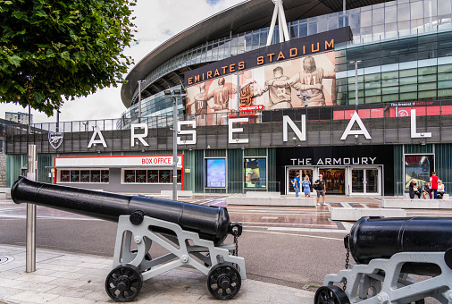 London, UK - People outside the exterior of Emirates Stadium, with canons, the emblem of Arsenal Football Club, in the foreground.