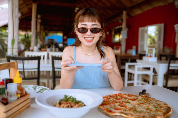 Woman using smart phone photographing pizza at restaurant. Asian customer using her mobile phone photographing pepperoni pizza for share on social media. fusion food stock pictures, royalty-free photos & images
