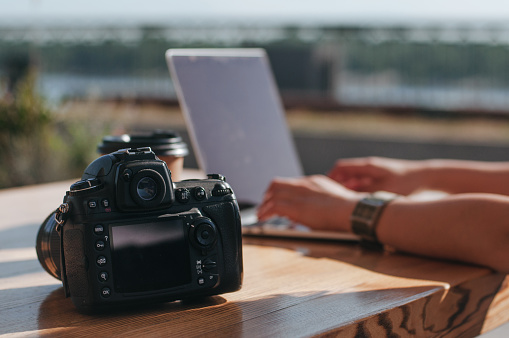 Camera on the table near female freelancer working on the laptop in the cafe outdoor. Freelance work concept. Selective focus on the camera.