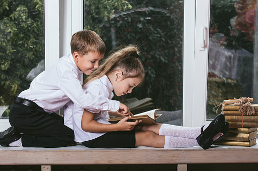 Happy boy and girl sitting on the windowsill reading books on the background of a window into a blooming garden