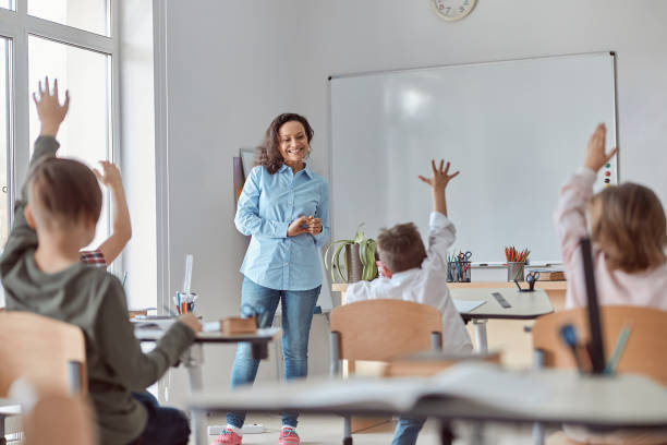 istock Young happy elementary school pupils at light modern classroom are rising their hands on a lesson 1332359746