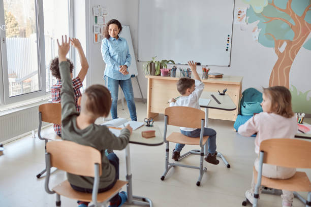 istock Young happy elementary school pupils at light modern classroom are rising their hands on a lesson 1332359751