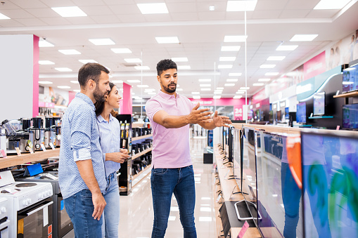 Couple seen in a home appliances store talking to a salesman in front of a TV set that they will buy for their new home.