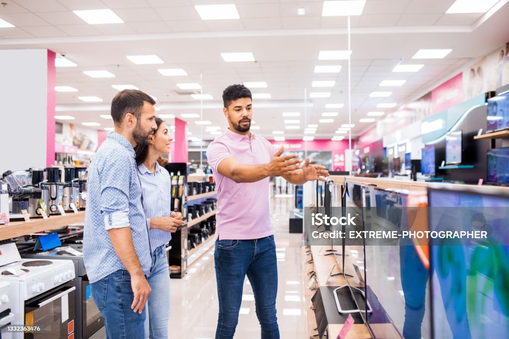 Salesman assists to a couple that wants to choose the best TV Couple seen in a home appliances store talking to a salesman in front of a TV set that they will buy for their new home. Television Set Stock Photo
