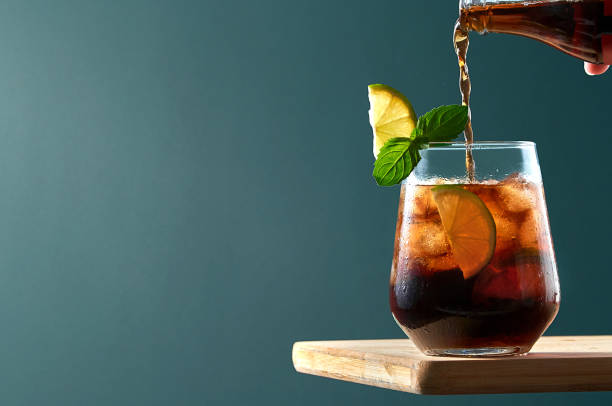 cuba libre or long island cocktail with pouring cola, rum and lime in glass on wooden board ob dark blue background. - 蘭姆酒 個照片及圖片檔