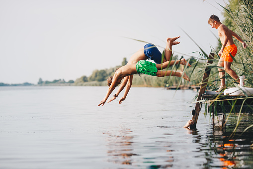 Three boys diving into river, jumping from jetty. They were out of water for few minutes and now all three of them are leaping. Around them is beautiful nature.