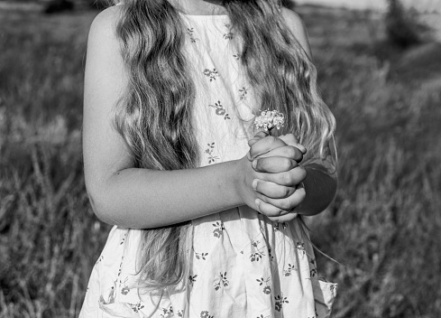 Adorable cheerful little girl in white cotton dress with flower in hand wild grass field. Happy child with long blond hair walking in countryside field. Outdoor rural road trip.
