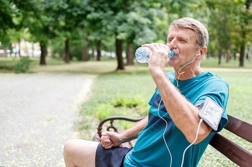 Older man siting on a park bench listening music and drinking water after a run