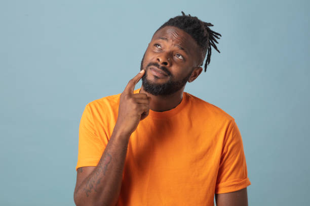Portrait of male wearing orange t-shirt looking at his chin with hand. Thoughtful studio portrait against blue background stock photo