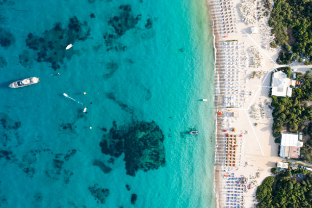 View from above, stunning aerial view of a white sand beach bathed by a turquoise water and relaxed people under some beach umbrellas at sunset. Cala di volpe beach, Costa Smeralda, Sardinia, Italy. View from above, stunning aerial view of a white sand beach bathed by a turquoise water and relaxed people under some beach umbrellas at sunset. Cala di volpe beach, Costa Smeralda, Sardinia, Italy. Cala Di Volpe stock pictures, royalty-free photos & images