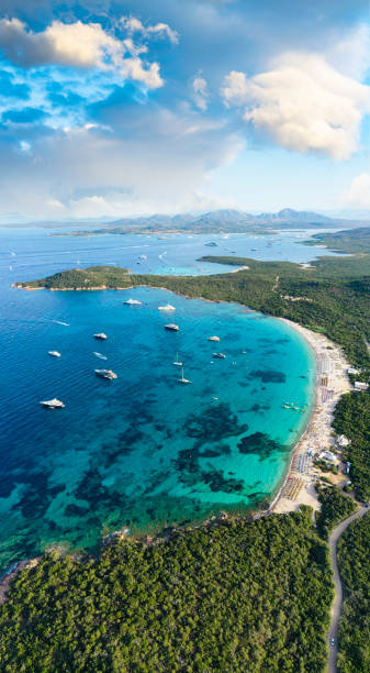 View from above, stunning aerial view of a green coastline with a white sand beach and and boats sailing on a turquoise water at sunset. Cala di volpe beach, Costa Smeralda, Sardinia, Italy. View from above, stunning aerial view of a green coastline with a white sand beach and and boats sailing on a turquoise water at sunset. Cala di volpe beach, Costa Smeralda, Sardinia, Italy. Cala Di Volpe stock pictures, royalty-free photos & images