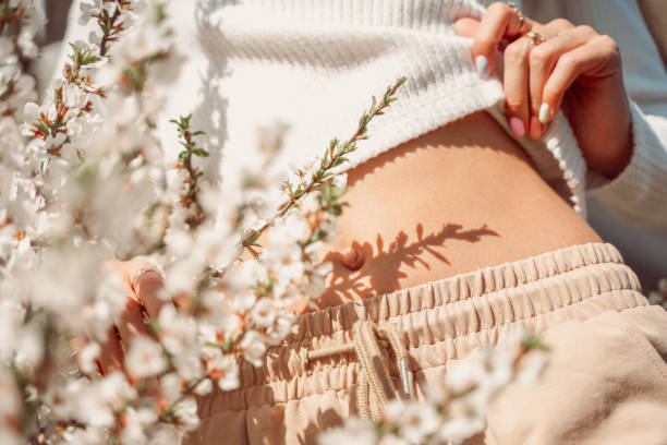 vue partielle de belle femme élancée dans des vêtements délicats posant avec les mains près de son ventre et avec des fleurs blanches en fleurs photo délicate, couleurs pastel. - bady care photos et images de collection