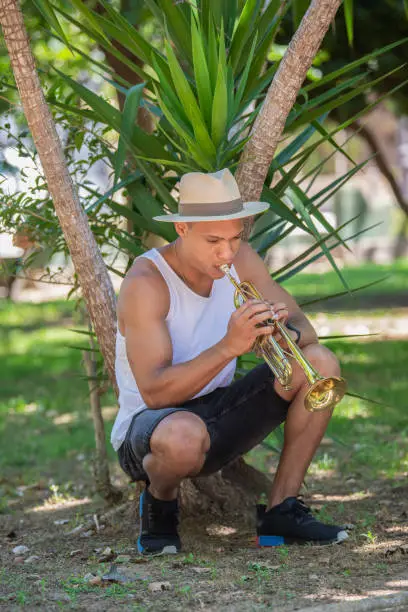 Photo of Latino man playing the trumpet crouched near a plant outdoors