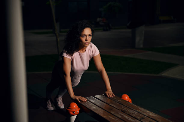 jeune athlète de nationalité mixte faisant des pompes à partir d’un banc en bois sur le terrain de sport et pompant les muscles de la poitrine et des épaules pendant l’entraînement du soir - athlete push ups muscular build female photos et images de collection