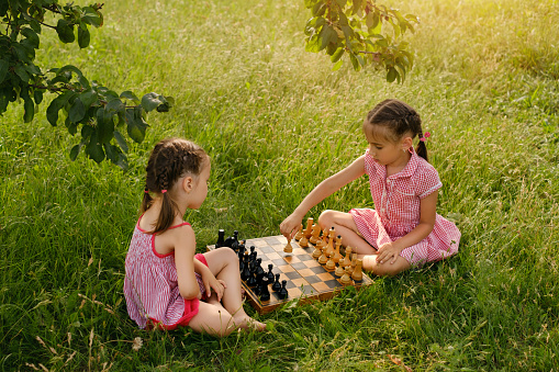 Two little girls game chess in the park in nature under a tree. An old wooden chessboard. Board intellectual educational games. Play outdoors. Unity with nature. Fresh air. Development of thinking