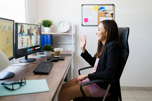 Team work meeting. Happy young woman wearing pajamas and a blazer waving to her co-workers during an online video call from home