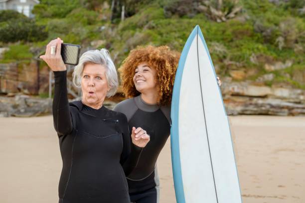 A vibrant senior woman and her mixed race granddaughter pose and take a selfie while spending the day together at the beach. The younger woman is holding a surfboard. The older woman is making a silly face. Both women are wearing wetsuits and are excited to spend the day surfing.