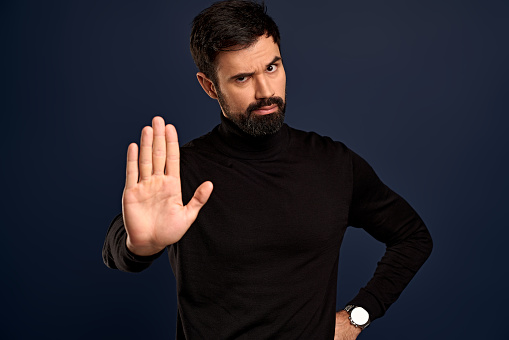 Judgemental and displeased, serious-looking handsome bearded man, grimacing from disapproval, dont permit something, raise arm in stop, never, prohibition gesture, standing Pacific Blue background.