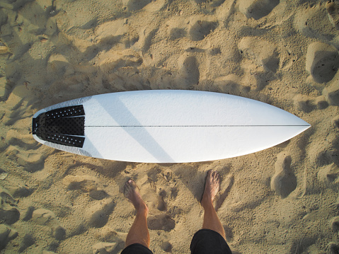 POV view of a man stands next white surfboard on the sandy beach. Surfing concept.