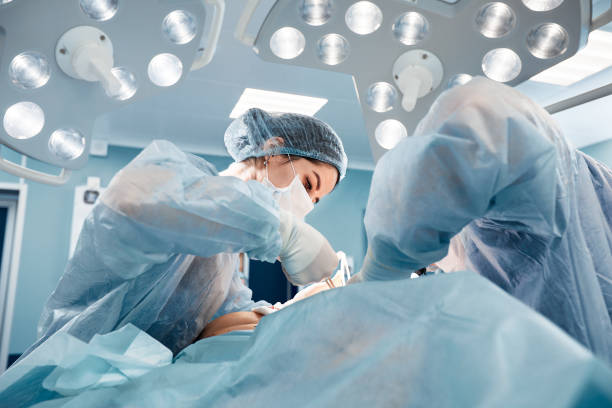 chirurghi uomini e donne in un'unica squadra durante un'operazione sul tavolo operatorio, i medici in attrezzature moderne eseguono un'operazione oncologica - chirurgo foto e immagini stock