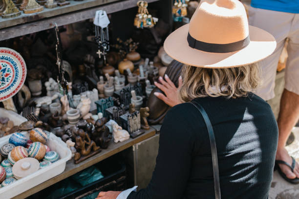 A delighted young woman examines a pink hat amongst an array of fashionable hats and colorful clothing at an outdoor market. High quality photo