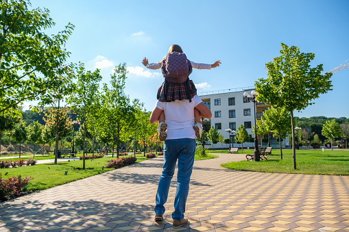 Daughter in school uniform and with backpack sitting on her father's shoulders with outstretched arms. Dad giving daughter piggyback ride when going to school, rear view
