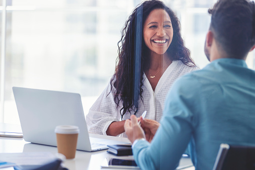 Business colleagues having a conversation. They are both young business people casually dressed in a bright office. Could be an interview or consultant working with a client. She is laughing and having fun. There is a laptop computer on the table  One person has his back to us.