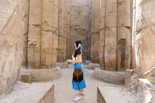 Side view of faceless stylish female traveler walking in between stone construction and admiring historic architecture in ancient ruined heritage building with hieroglyphs on weathered columns. Trip concept