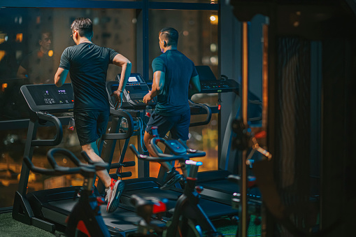 rear view body macho men running treadmill together in gym at night facing city street light