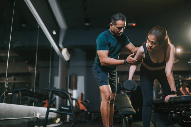 istruttore di fitness personal trainer che guida la donna asiatica di mezza età che si allena con manubri in posizione affondo sulla panchina della palestra di notte - women weight bench exercising weightlifting foto e immagini stock