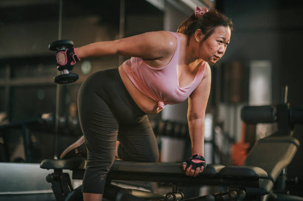 夜にジムのベンチで突進位置にダンベルで運動する体陽性アジアの中年女性 - gym weight bench exercising weights ストックフォトと画像