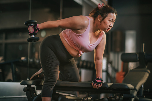 body positive Asian mid adult woman exercising with dumbbells in a lunge position at gym bench at night
