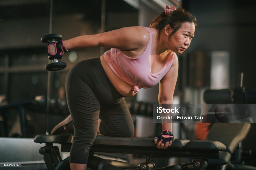 body positive Asian mid adult woman exercising with dumbbells in a lunge position at gym bench at night body positive Asian mid adult woman exercising with dumbbells in a lunge position at gym at night One Woman Only Stock Photo