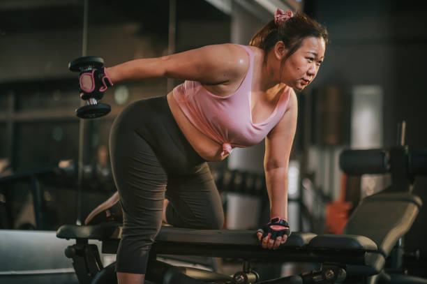 body positive Asian mid adult woman exercising with dumbbells in a lunge position at gym bench at night body positive Asian mid adult woman exercising with dumbbells in a lunge position at gym at night exercise machine photos stock pictures, royalty-free photos & images