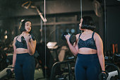body positive Asian mixed race teenage girl carrying dumbbells looking at mirror reflection in gym