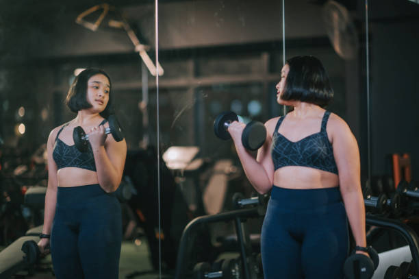 body positive Asian mixed race teenage girl carrying dumbbells looking at mirror reflection in gym body positive Asian mixed race teenage girl carrying dumbbells looking at mirror reflection in gym body confidence stock pictures, royalty-free photos & images