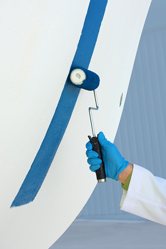 Closeup man in boatyard marina with a gloved hand painting a large boat with antifouling paint with a roller giving a protective coating to keep barnacles off the hull bottom.