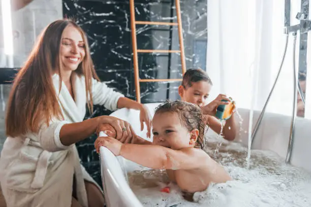 Photo of Young mother helps her son and daughter. Two kids washing in the bath