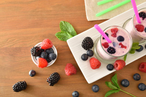 Forest fruit smoothie with milk in glass glasses on white plate and fruits on wooden bench. Top view.