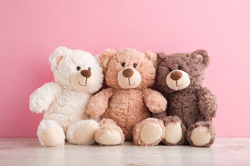 Smiling white, light brown and dark brown teddy bears sitting on table at pink wall background. Pastel color. Togetherness and friendship concept. Front view. Closeup.