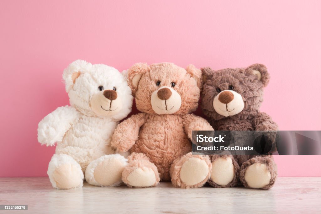 Smiling white, light brown and dark brown teddy bears sitting on table at pink wall background. Pastel color. Togetherness and friendship concept. Front view. Closeup. Teddy Bear Stock Photo