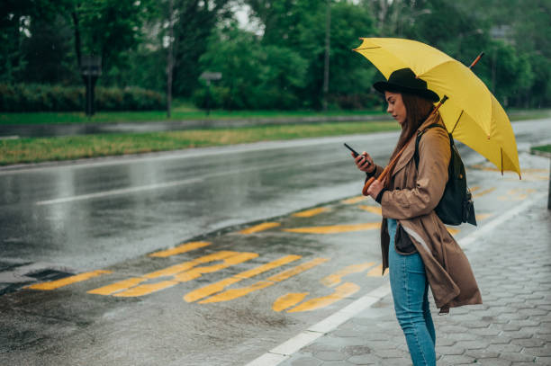 istock Woman using a smartphone and holding a yellow umbrella while waiting for a public transport 1332457871