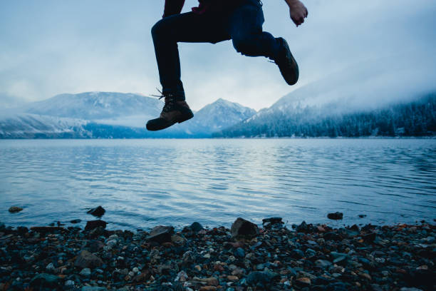 An unrecognizable person jumping above a cold winter lake with snow capped mountains in the background. They are wearing hiking boots and a plaid shirt.