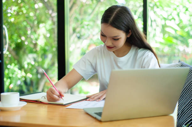 istock Work from home concept. Young woman writing notebook while working on laptop at home. 1332464064