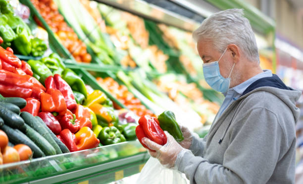 Senior adult man with surgical mask due to coronavirus while shopping at supermarket. White-haired retiree looking at red and green peppers in the vegetable department. Consumerism concept Senior adult man with surgical mask due to coronavirus while shopping at supermarket. White-haired retiree looking at red and green peppers in the vegetable department. Consumerism concept vegetable stand stock pictures, royalty-free photos & images